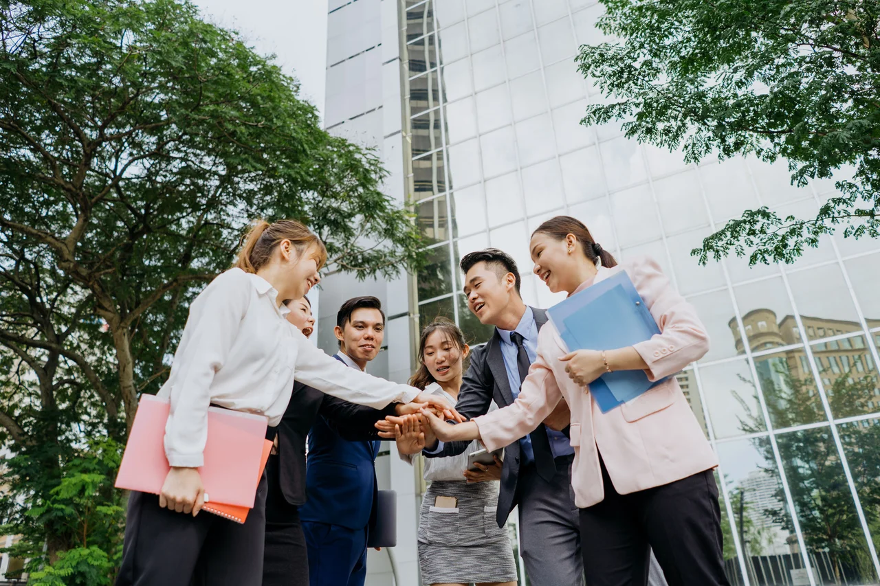 Group of professional colleagues stacking their hands together in front of a modern glass office building.