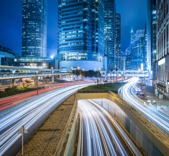 夜間城市多線道道路上車流光軌的長曝光照片，光軌穿過照明璀璨的摩天大樓群並延伸至隧道入口。