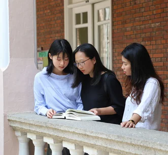 三名女子在設有白色欄杆及紅磚牆的戶外走廊旁一同閱讀書籍。
