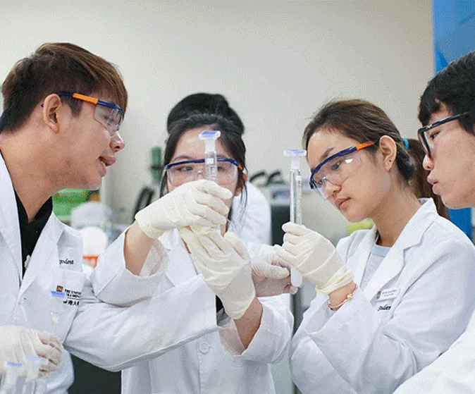 身穿白袍、佩戴防護眼鏡及手套的港大學生在實驗室內一同檢視試管。