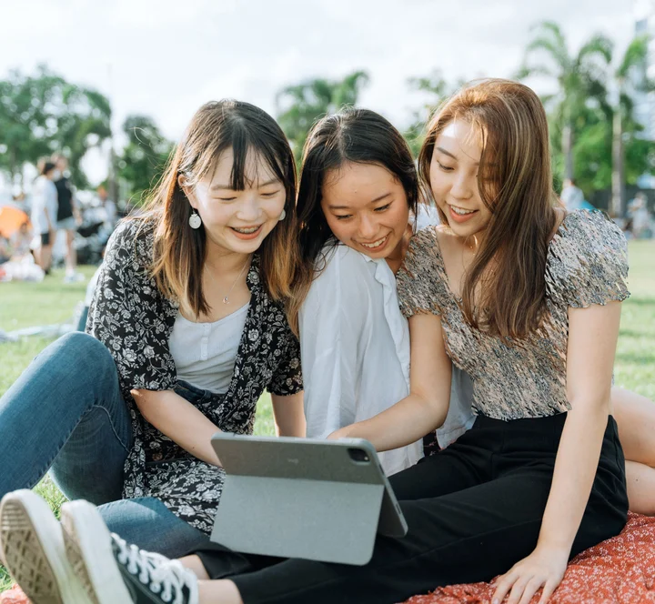三名笑容滿面的年輕女子坐在戶外草坪上，一同看著平板電腦。