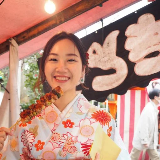 一名身穿花卉圖案和服的年輕女子在戶外集市攤位前微笑，手中拿著一串烤肉。