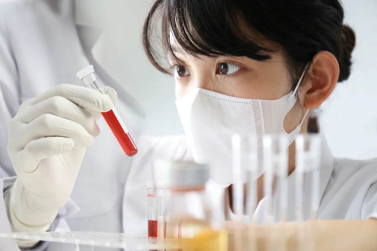一名佩戴口罩和手套的研究人員在實驗室內仔細觀察盛有紅色液體樣本的試管。